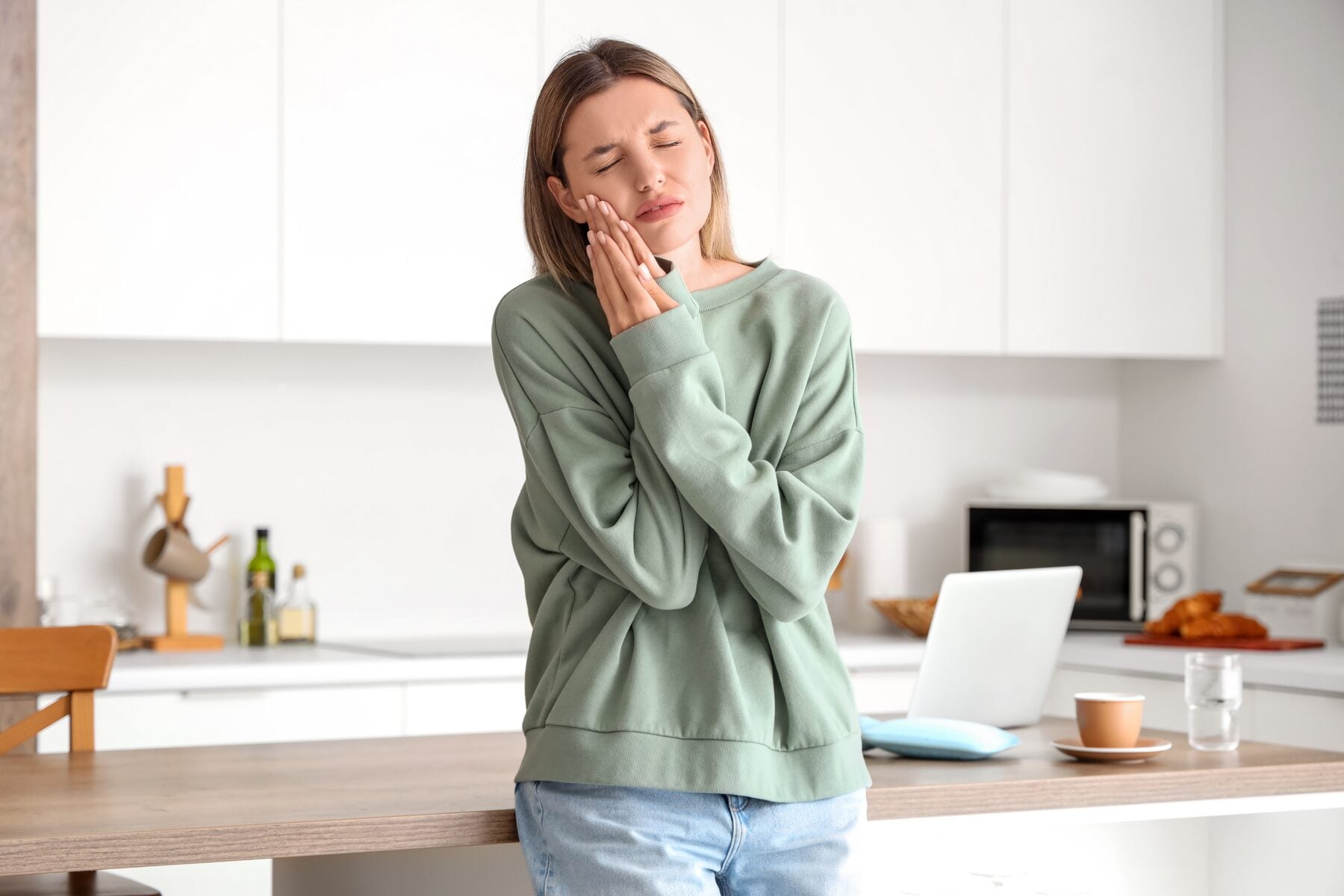 A woman holding her jaw in discomfort in her kitchen